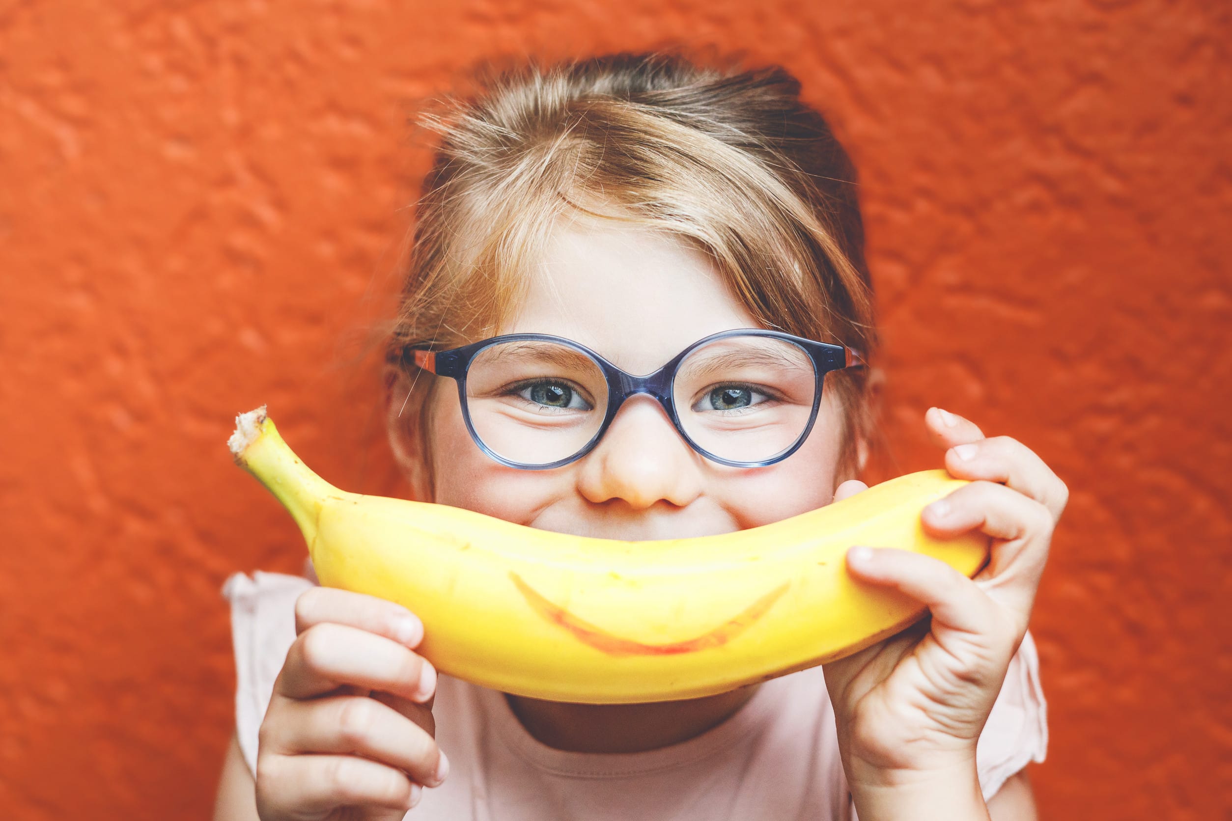 Happy little child girl with yellow banana like smile on orange background. Preschool girl with glasses smiling. Healthy fruits for children snacks marketed to children