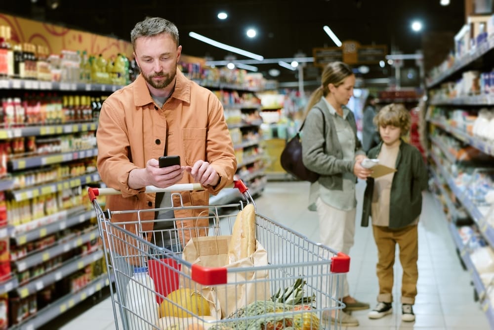 Young,Man,Checking,Phone,While,Pushing,Shopping,Cart,In,Grocery grocery store